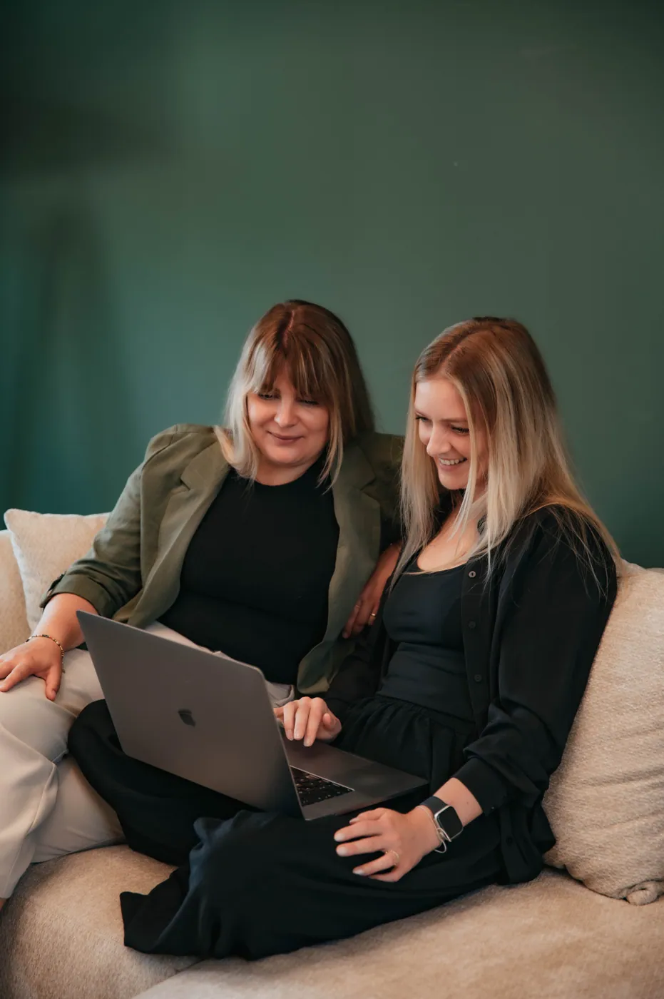 Zwei Frauen arbeiten lachend gemeinsam am Laptop auf einem Sofa